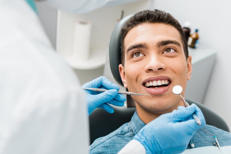 A man is seated in a dental chair, looking up as a dentist, whose hands are visible and gloved, examines his teeth with dental tools.