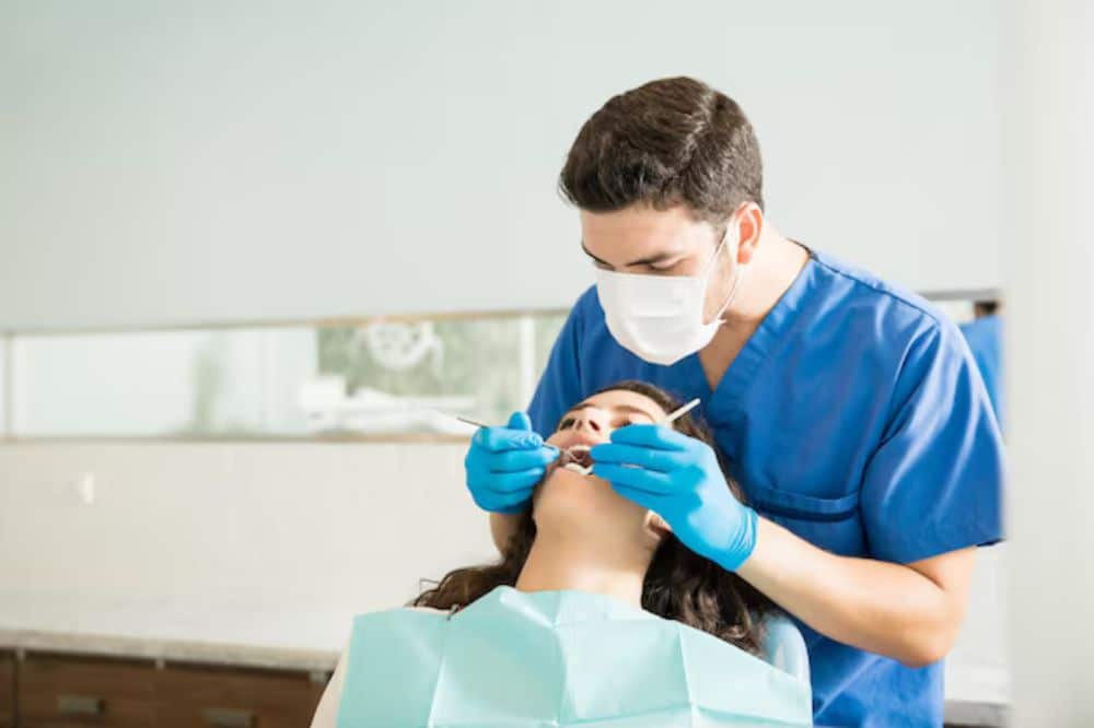 A male dentist wearing a face mask and blue scrubs is performing a checkup or dental procedure on a female patient who is reclined in the exam chair.