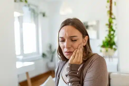 A concerned woman with her eyes closed is holding her cheek and jaw with her hand, clearly experiencing pain or discomfort from a toothache or earache in a bright, modern living room.