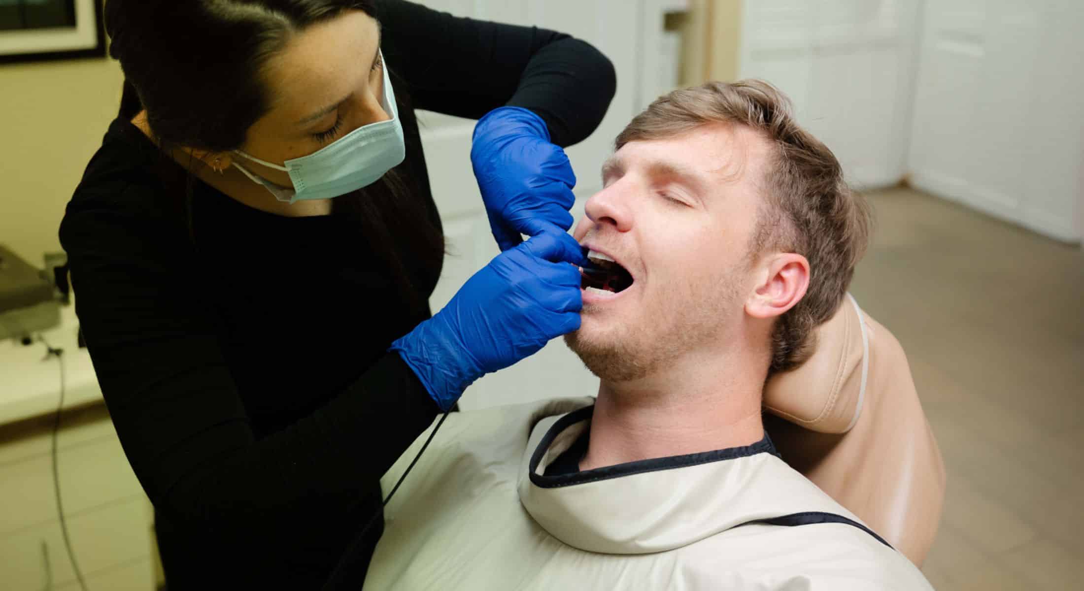 Dentist wearing gloves and a face mask examining a patient's mouth while the patient reclines in a dental chair.