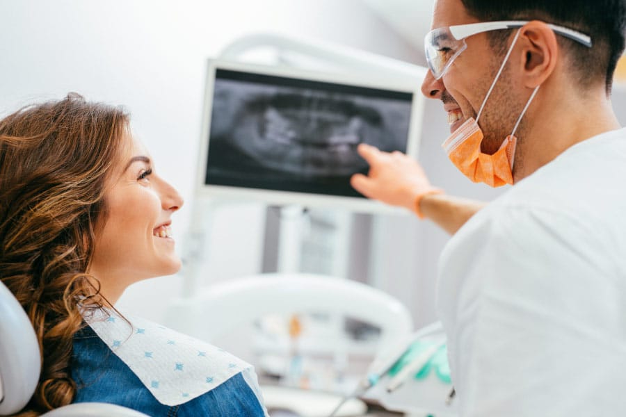 Dentist pointing to a dental panoramic X-ray on a monitor while discussing the results with a smiling patient.